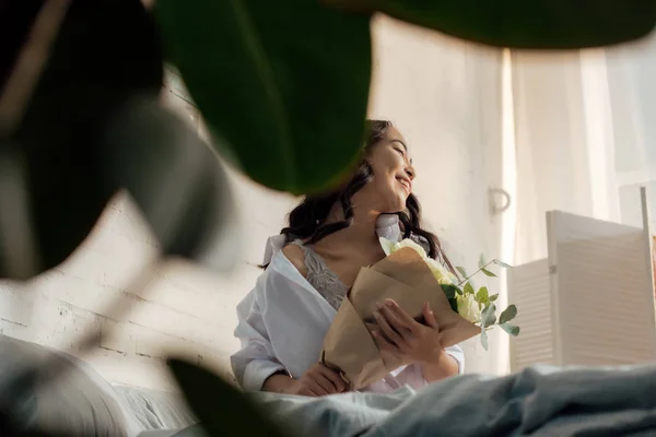 Foco seletivo de bela feliz jovem mulher asiática em camisa branca sentado na cama com buquê de rosas — Fotografia de Stock