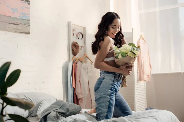 Happy attractive asian girl kneeling on bed and holding beautiful flower bouquet — Stock Photo
