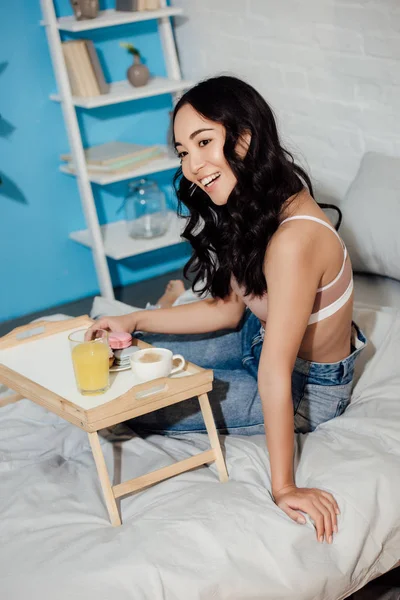 Hermosa feliz asiático chica sentado en cama y disfrutar de desayuno en casa - foto de stock