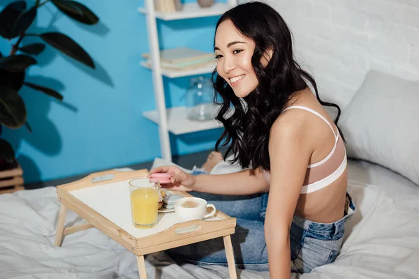 Atractiva mujer asiática sentada en la cama y disfrutando del desayuno en casa - foto de stock