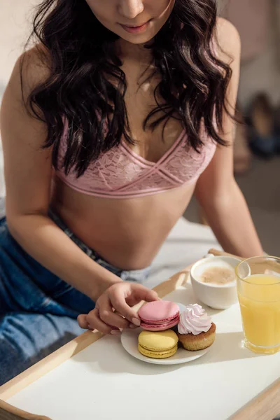 Recortado disparo de sensual joven mujer en sujetador comer macarrones para el desayuno en el dormitorio - foto de stock