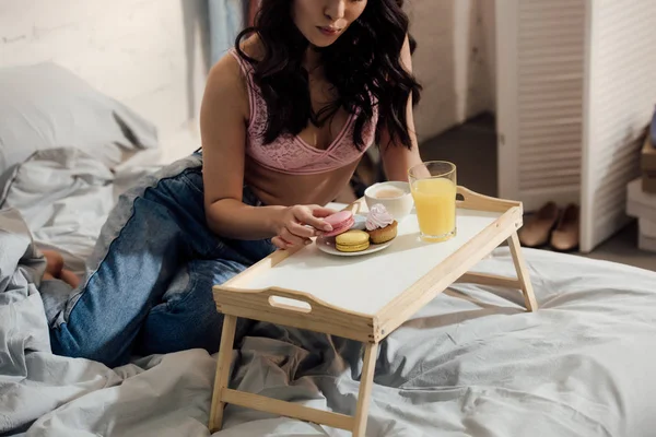 Tiro cortado de menina atraente segurando macaron saboroso enquanto toma café da manhã no quarto — Fotografia de Stock