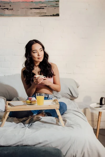 Attractive asian girl holding cup of coffee and having breakfast in bedroom — Stock Photo