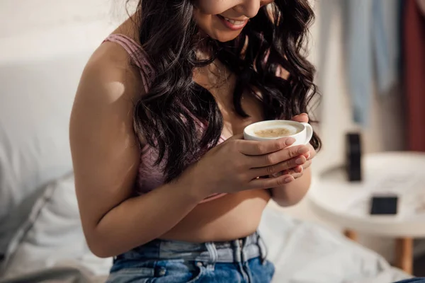 Recortado disparo de sonriente mujer joven en sujetador sosteniendo taza de capuchino en el dormitorio - foto de stock