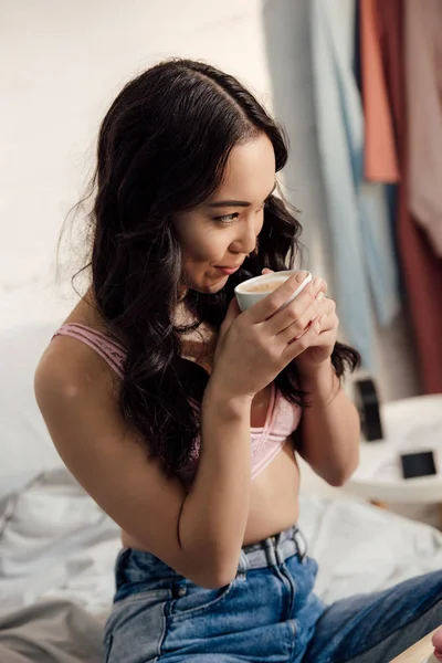 Hermosa sonriente chica asiática en sujetador beber café en dormitorio - foto de stock