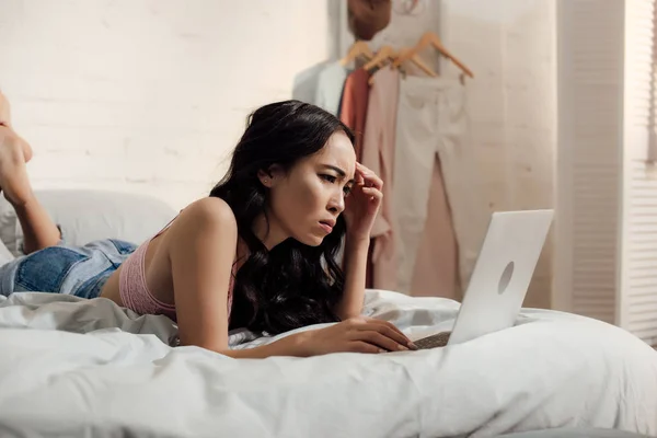 Серьезная хмурая молодая женщина, использующая ноутбук в постели — стоковое фото