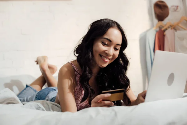 Hermosa sonrisa chica asiática celebración de tarjeta de crédito y el uso de ordenador portátil en el dormitorio - foto de stock
