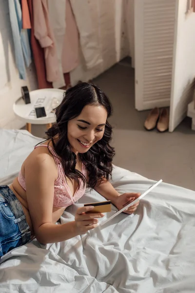 Vista de ángulo alto de la joven feliz de compras en línea con tableta digital y tarjeta de crédito en el dormitorio - foto de stock