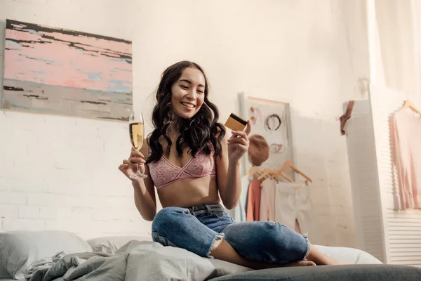 Красивая улыбающаяся азиатка с бокалом вина и кредитной картой в спальне — стоковое фото