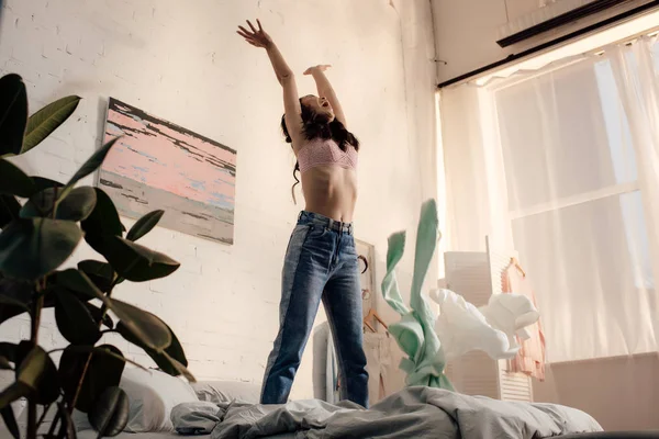 Низкий угол обзора эмоциональной молодой женщины, стоящей на кровати и поднимающей руки — стоковое фото
