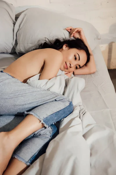 Красивая улыбающаяся молодая азиатка в джинсовых штанах лежит на кровати та дома — стоковое фото