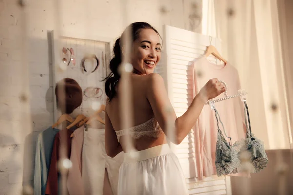 Beautiful asian girl holding hanger with lace bralette and smiling at camera — Stock Photo