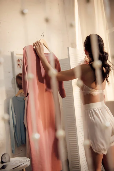 Back view of young woman in underwear holding hanger with fashionable dress and looking away in wardrobe — Stock Photo