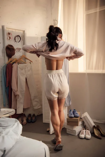 Vista posterior de la mujer joven que se pone el suéter elegante y elegir el calzado en casa - foto de stock