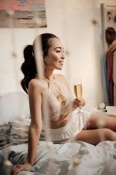 Bela sorrindo asiático menina no cueca sentado no cama com copo de champanhe e olhando para longe — Fotografia de Stock