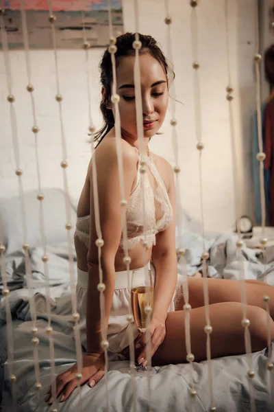 Sedutor jovem asiático mulher sentado no cama com copo de champanhe — Fotografia de Stock