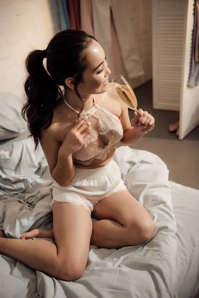 Vista ad alto angolo di bella ragazza sorridente in lingerie che tiene un bicchiere di champagne in camera da letto — Foto stock