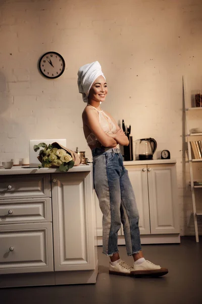 Красивая улыбающаяся азиатская девушка в кружевном бюстгальтере и полотенце на голове стоя со скрещенными руками и глядя на дом — стоковое фото