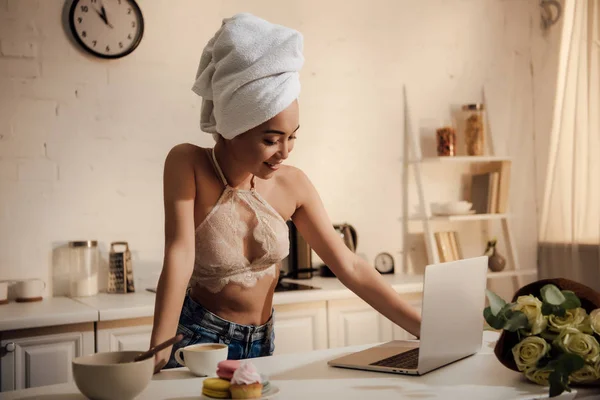 Schöne lächelnde junge Frau in Spitzen-Bralette und Handtuch auf dem Kopf mit Laptop beim Frühstück zu Hause — Stockfoto