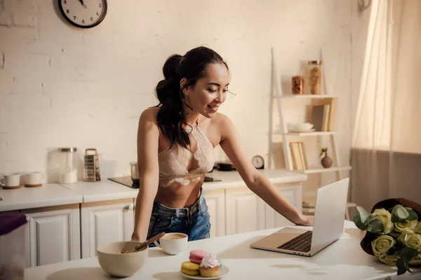 Красивая улыбающаяся молодая женщина в кружевном бюстгальтере и джинсах с ноутбуком во время завтрака дома — стоковое фото