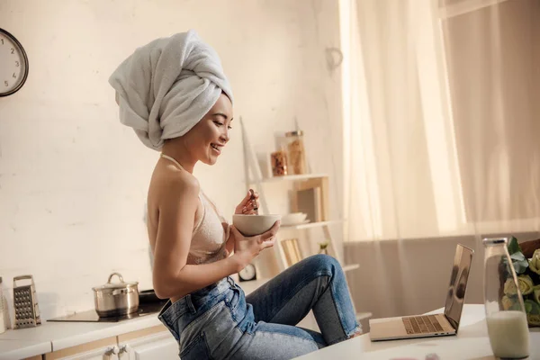 Улыбающаяся девушка в кружеве бюстгальтер и полотенце на голове едят кукурузные хлопья на завтрак и с помощью ноутбука — стоковое фото