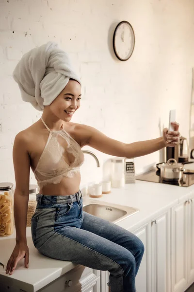 Привлекательная улыбающаяся молодая женщина с полотенцем на голове делает селфи со смартфоном на кухне — стоковое фото