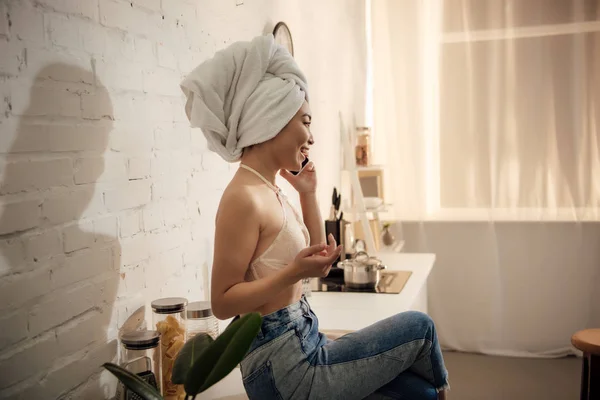 Side view of attractive asian girl with towel on head talking by smartphone while sitting in kitchen — Stock Photo