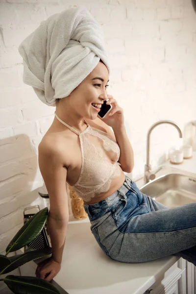 Привлекательная азиатка с полотенцем на голове разговаривает по смартфону, сидя на кухне — стоковое фото