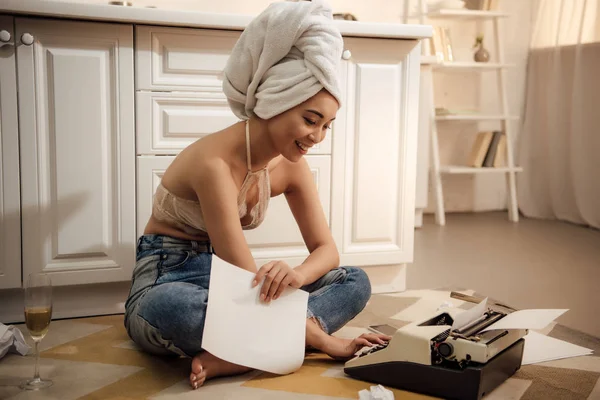 Smiling young woman with towel on head holding paper and using typewriter while sitting on floor — Stock Photo
