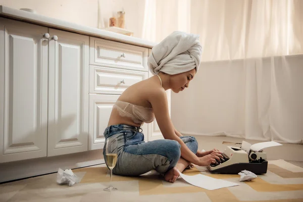 Vista laterale di attraente donna asiatica con asciugamano sulla testa seduta nel pavimento e utilizzando macchina da scrivere — Foto stock