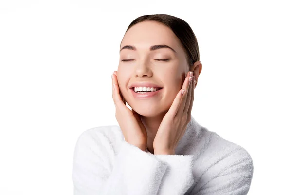 Hermosa mujer sonriente en albornoz con los ojos cerrados tocando la cara aislada en blanco - foto de stock