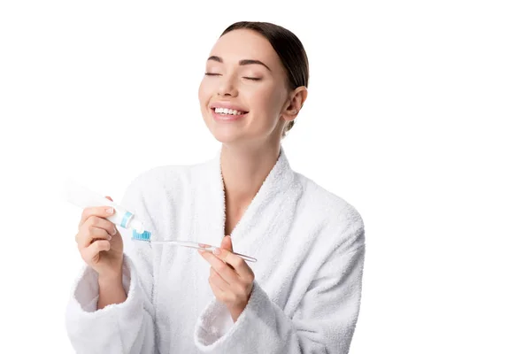 Beautiful happy woman in white bathrobe putting toothpaste on toothbrush isolated on white — Stock Photo
