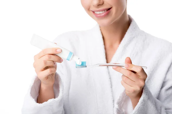 Vista parcial de la mujer en albornoz blanco poniendo pasta de dientes en el cepillo de dientes aislado en blanco - foto de stock