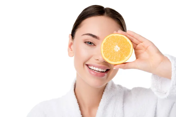 Hermosa mujer en albornoz blanco sosteniendo naranja en frente de la cara y mirando a la cámara aislada en blanco - foto de stock