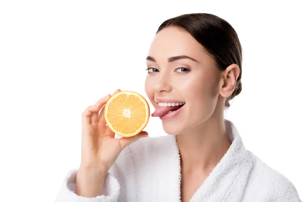 Mujer sonriente en albornoz blanco sosteniendo naranja y sacando la lengua aislada en blanco - foto de stock