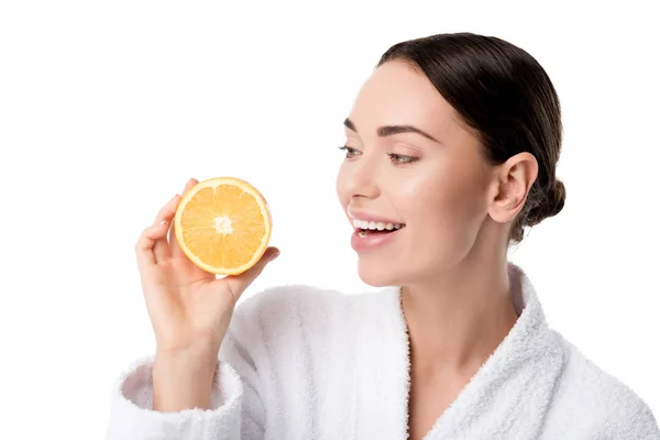 Hermosa mujer sonriente en albornoz blanco sosteniendo naranja aislado en blanco - foto de stock