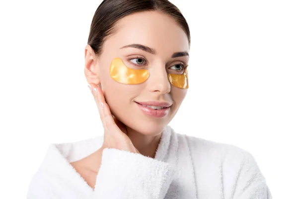 Hermosa mujer sonriente con parches de ojos dorados tocando la cara y mirando hacia otro lado aislado en blanco - foto de stock