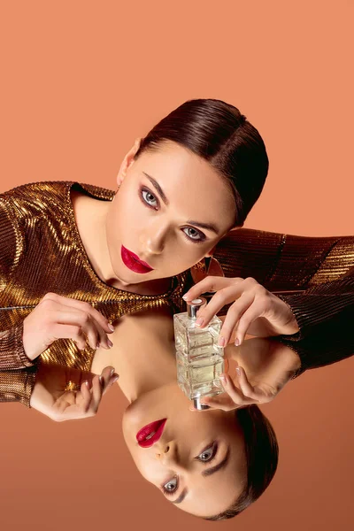 Attractive woman in golden clothes with glamorous makeup, perfume bottle and mirror reflection posing isolated on orange — Stock Photo