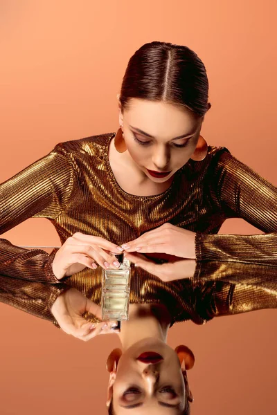 Hermosa mujer en ropa dorada con botella de perfume y espejo reflejo aislado en naranja - foto de stock