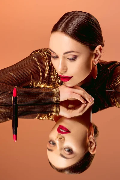 Atractiva mujer con maquillaje glamoroso, reflejo de espejo y lápiz labial rojo posando aislado en naranja - foto de stock