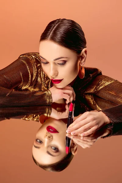 Hermosa mujer con glamoroso maquillaje sosteniendo lápiz labial rojo y posando con reflejo de espejo aislado en naranja - foto de stock