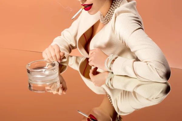 Vista cortada da mulher no colar de pérolas com espelho reflexão fumar cigarro isolado na laranja — Fotografia de Stock