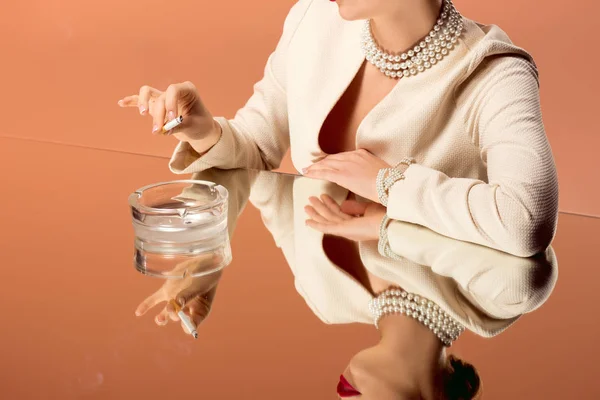 Vista recortada de la mujer en collar de perlas con espejo reflexión celebración de cigarrillo sobre cenicero aislado en naranja - foto de stock