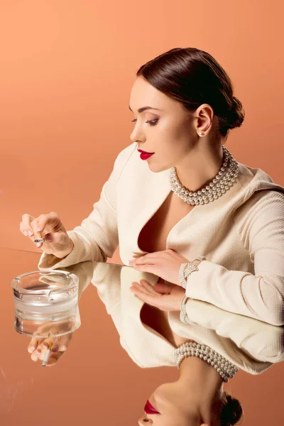 Hermosa mujer glamorosa en collar de perlas con espejo reflexión celebración de cigarrillo sobre cenicero aislado en naranja - foto de stock