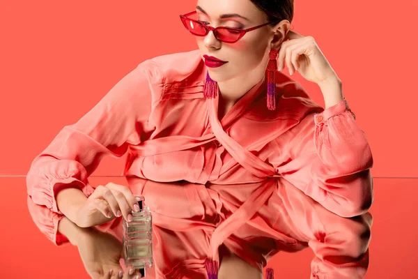 Mujer con estilo en blusa y gafas de sol rojas posando con botella de perfume y espejo reflejo aislado en coral vivo - foto de stock