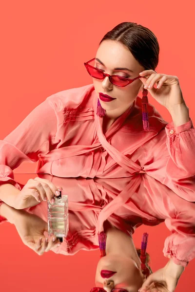 Mujer glamorosa en blusa y gafas de sol rojas posando con botella de perfume y espejo reflejo aislado en coral vivo - foto de stock