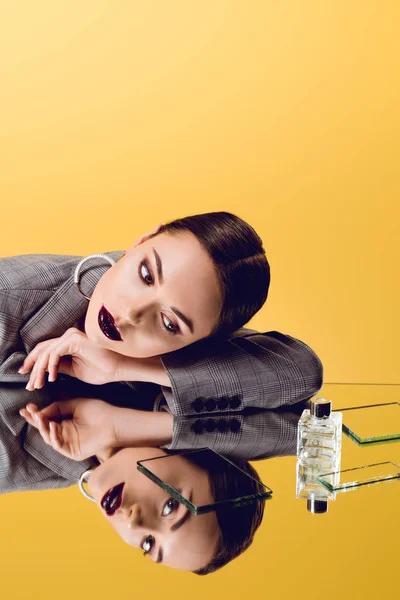 Atractiva mujer glamorosa en desgaste formal con reflejo de espejo y botella de perfume aislado en amarillo - foto de stock