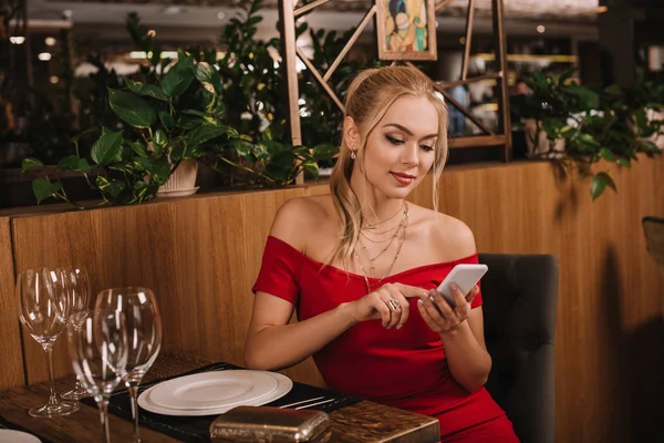 Attractive woman in red dress sitting in restaurant and using smartphone — Stock Photo