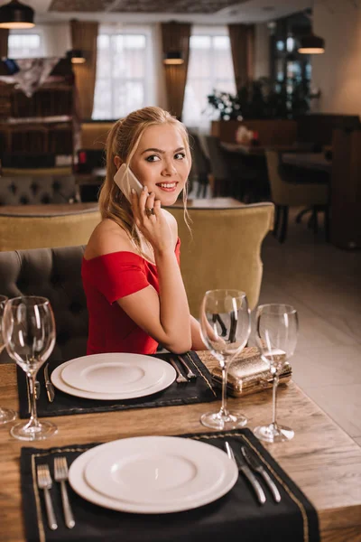 Mulher atraente em vestido vermelho sentado no restaurante usando smartphone e olhando para a câmera — Fotografia de Stock
