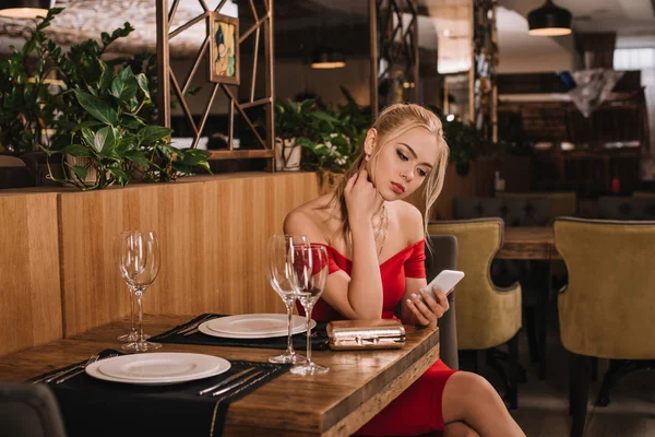 Attraktive Frau in rotem Kleid sitzt im Restaurant und schaut aufs Smartphone — Stockfoto